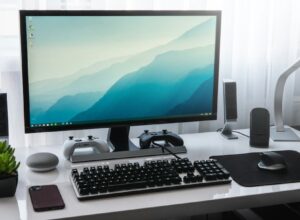 A computer monitor, keyboard, and game controllers on a white desk in a workspace