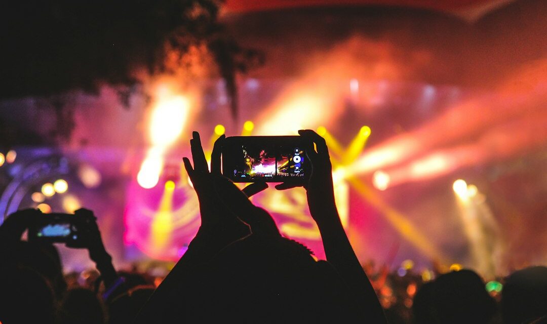Person holding smartphone taking video of a concert near stage with lights during nighttime